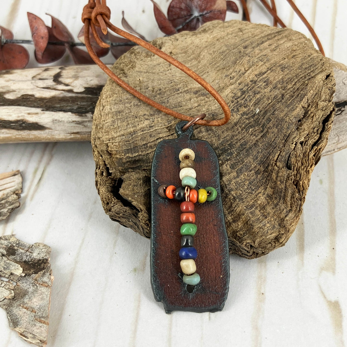 Beaded cross on aged metal pendant made by Linda Stiles Smith of Naturescapes Studio hung on light brown leather cords with knoted adjustable ends.