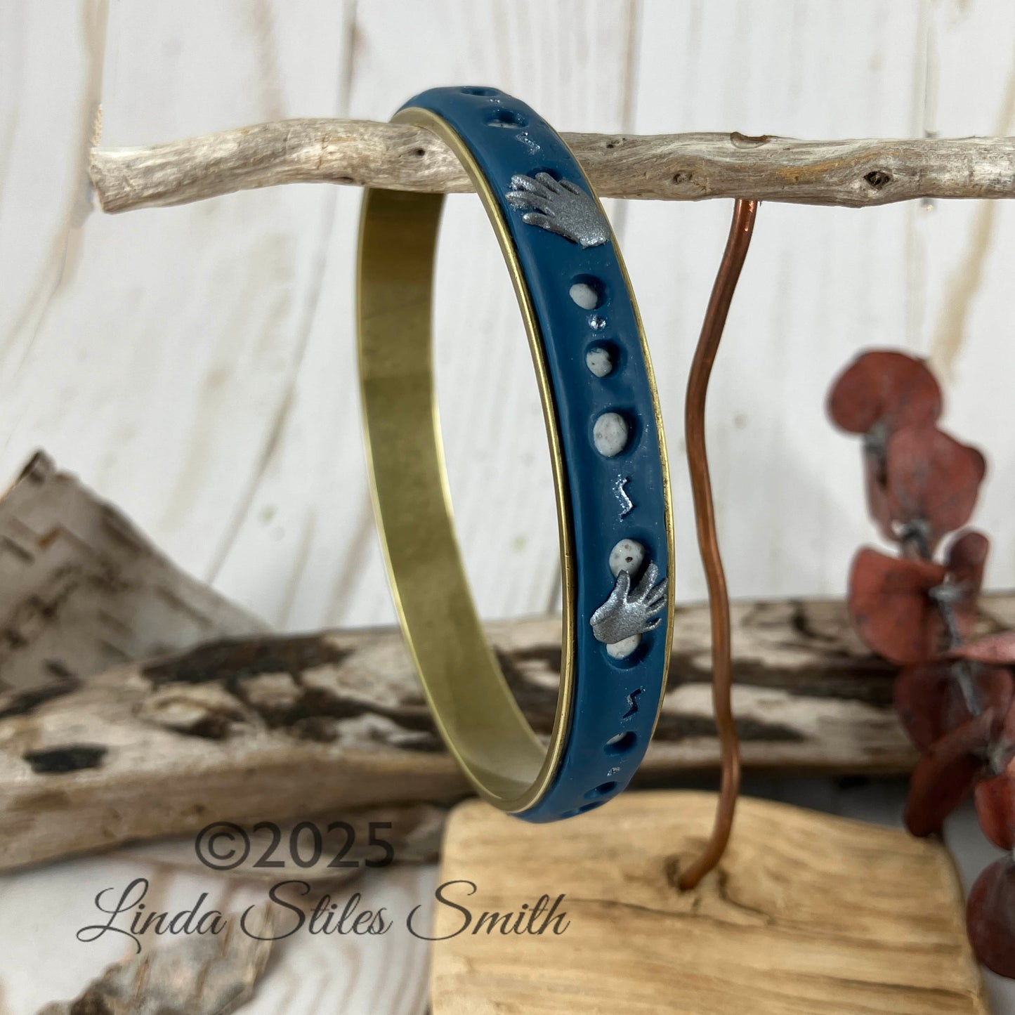 Bangle bracelet with brass covered in blue and grey polymer clay layers. Tiny silver hands on the surface among cut out round holes showing clay layered beneath. Handcrafted at NatureScapes Studio by Linda Stiles Smith