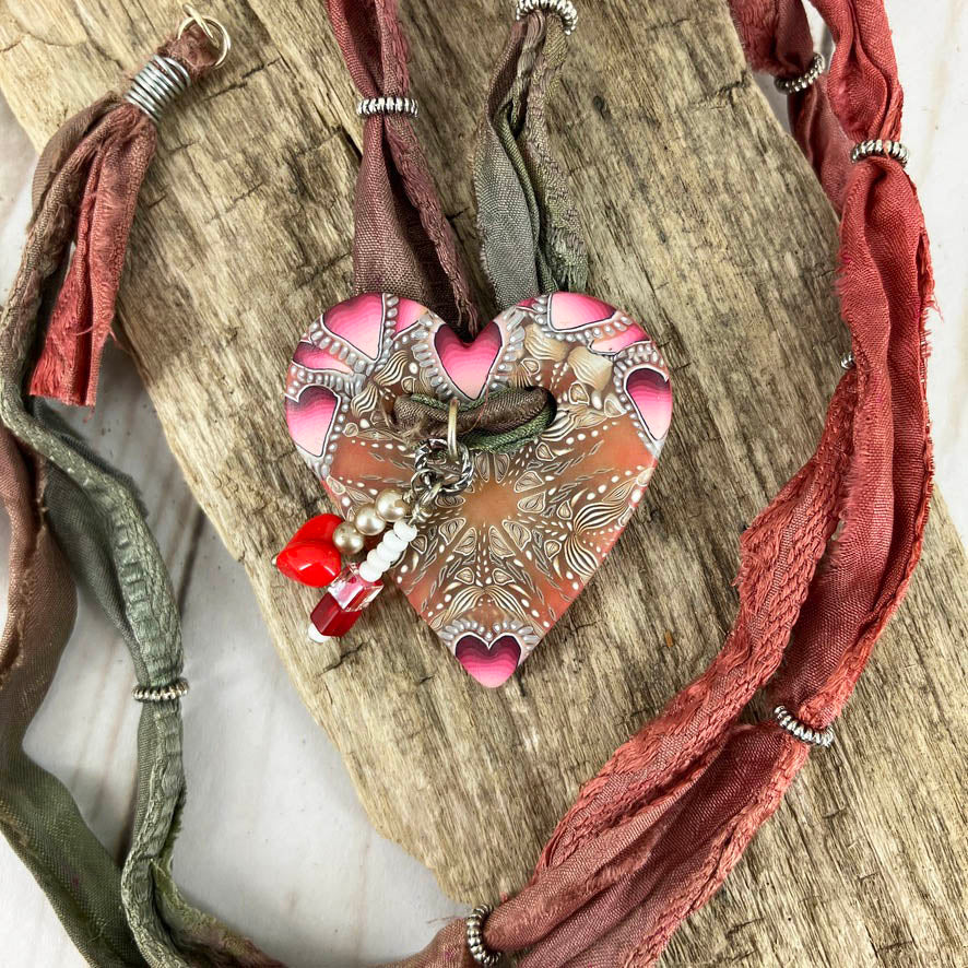 A red and caramel heart button becomes a necklace with a colorful sari silk cording all adorned with metal ring beads and a red heart dangle. A darling design by Linda Stiles Smith
