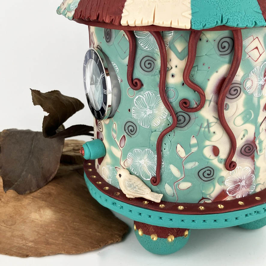 Tendrils of red clay drape down the side of the clock from the roof of the carousel clock almost touching the tiny white bird on the edge of the carousel clock.