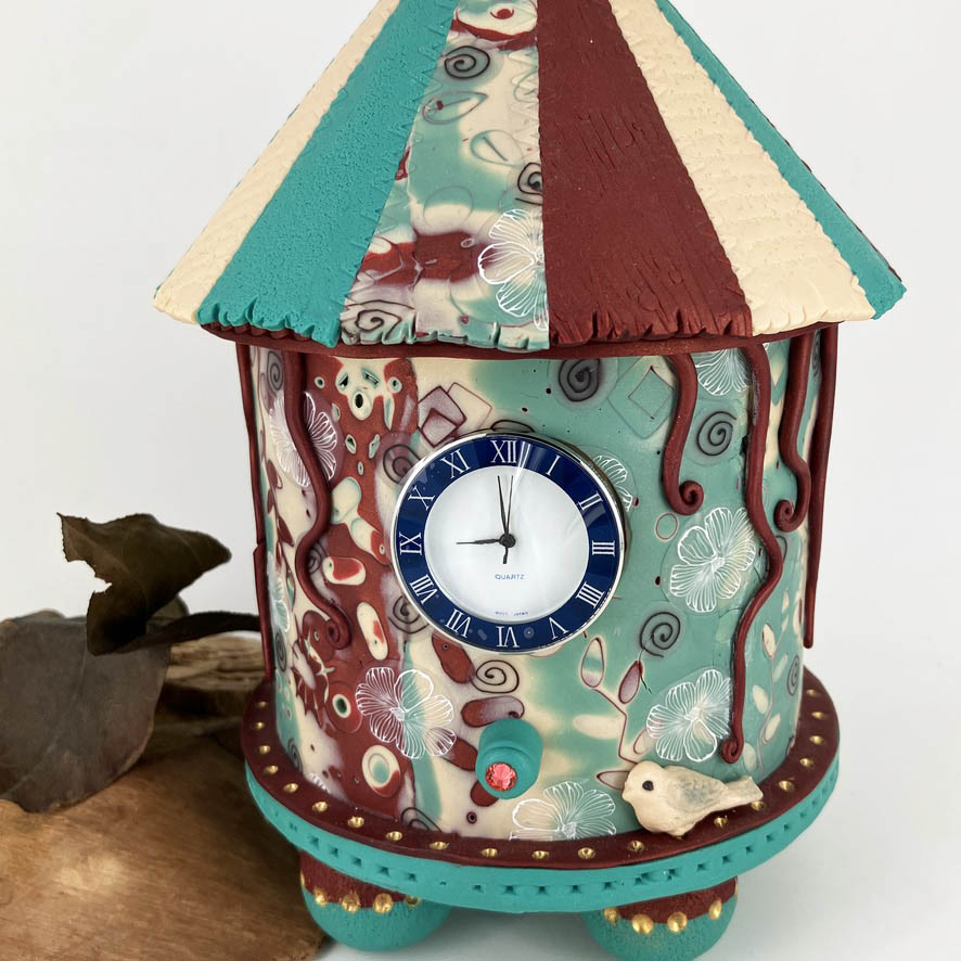 The quartz clock movement of the handcrafted carousel clock is nestled above the birds perch on the front of the turquoise, ivory and red patterned round form.