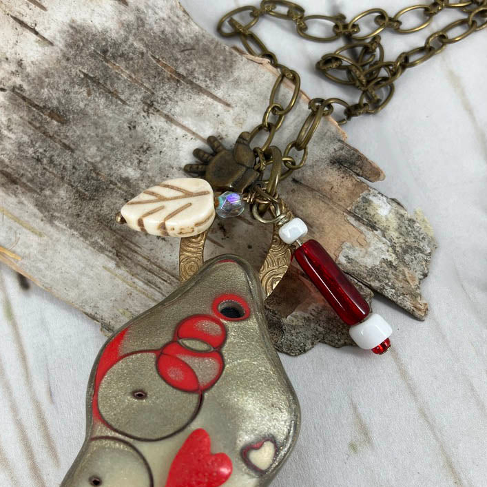 Special charms of the heart filled figure necklace sports a leaf, red glass bead and brass hand to accent the statement necklace handmade by Linda Stiles Smith