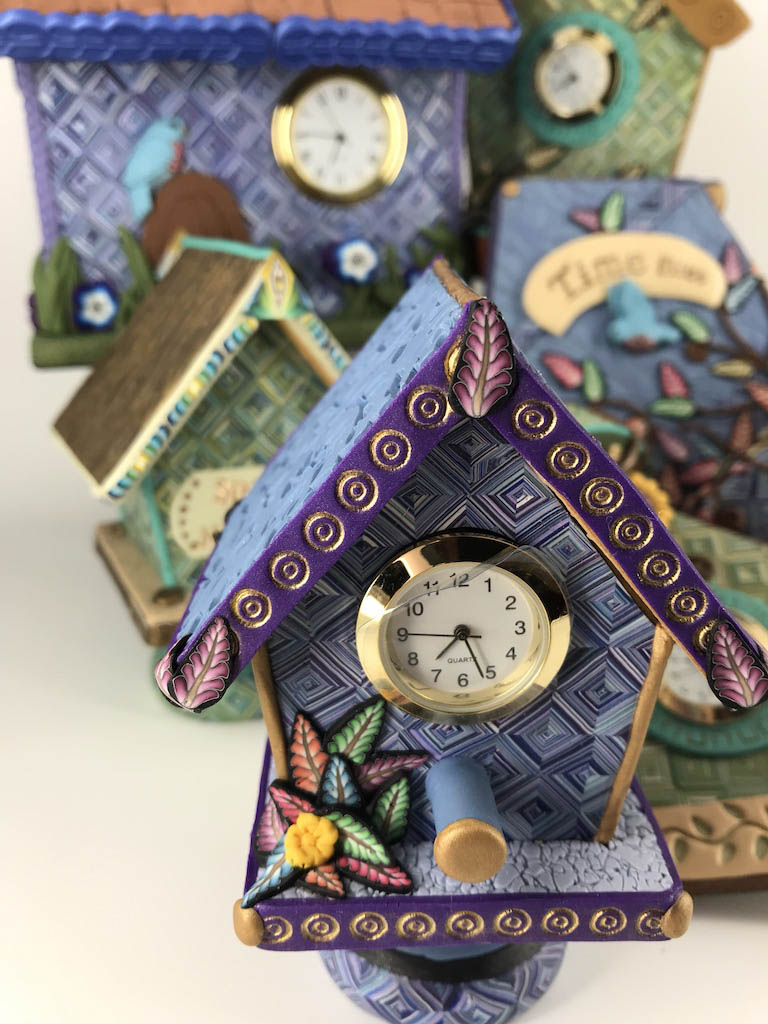 Time Flies Blue and Purple Bird House Desk Clock handmade with Polymer Clay by Linda Stiles Smith of NatureScapes Studio. Front view surrounded by other handmade clocks. Gift ideas for women.